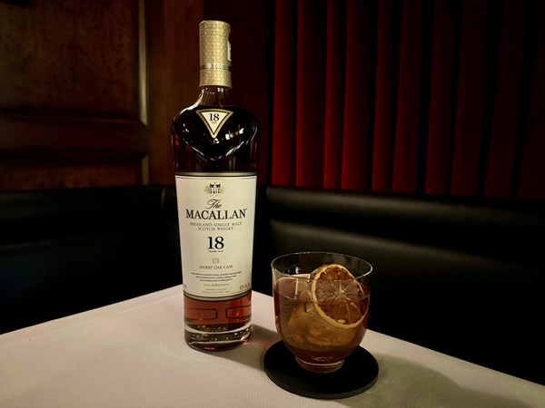 Bottle of The Macallan 18 year next to a whiskey cocktail with a drie lemo wedge, on a white table cloth